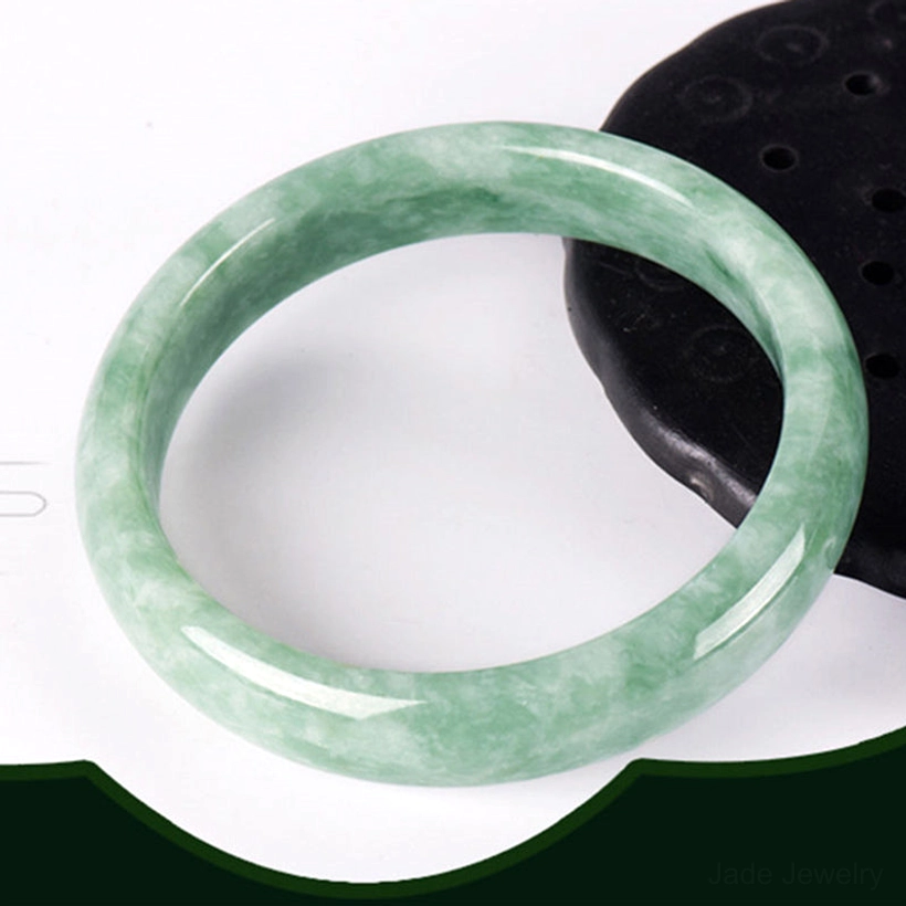 Natural jade bangle bracelet with muted green vein patterns photographed flat overhead on dark green backdrop with black stone props, highlighting authentic jade texture and color variations