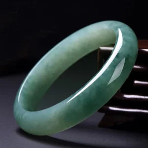 Natural jade bangle bracelet with deep green tone and subtle internal veining displayed on dark wooden surface, showing three-quarter view of polished craftsmanship