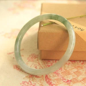 Handcrafted jade bangle bracelet showing mixed light and dark green jade texture with polished smooth surface on stacked kraft boxes