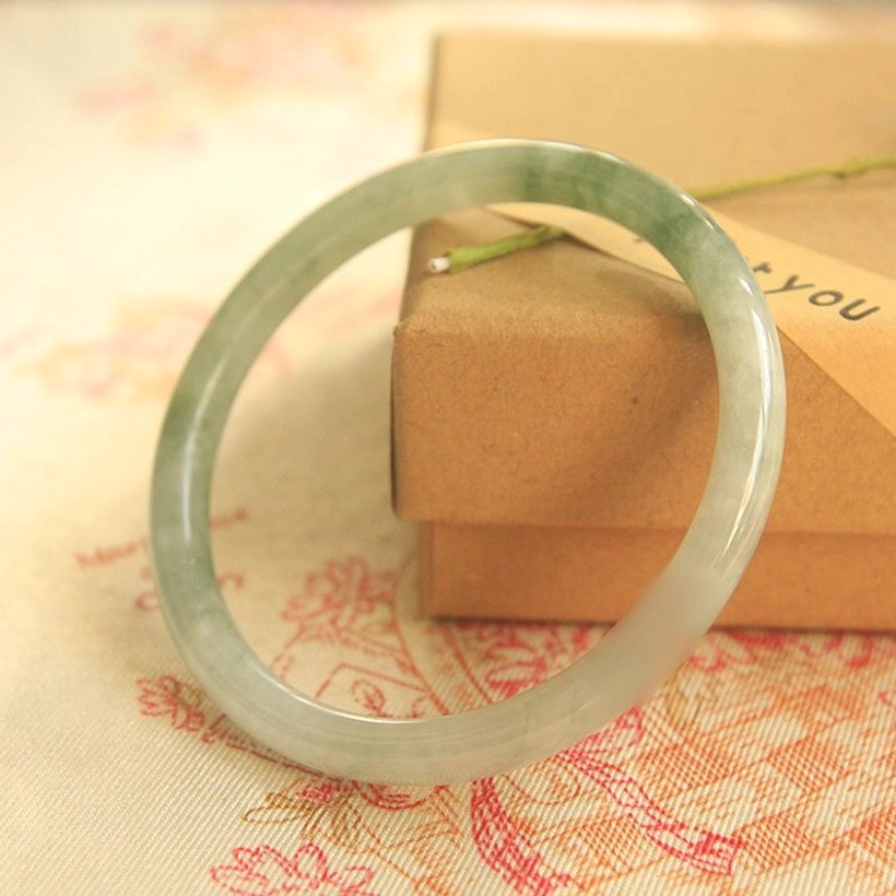 Handcrafted jade bangle bracelet showing mixed light and dark green jade texture with polished smooth surface on stacked kraft boxes