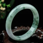 Natural jade bangle bracelet with dark green spotted texture, smooth polished finish, overhead view on wooden prop