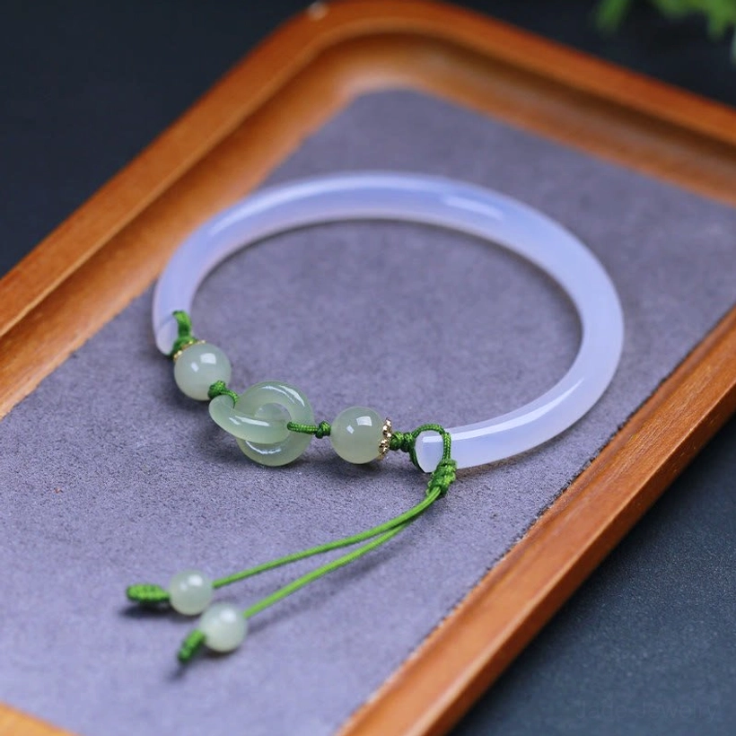Pale blue-white jade bangle with green braided cord and jade beads, side overhead view on wooden tray with purple velvet mat