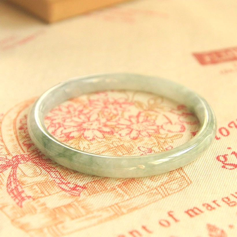 Slim round jade bangle bracelet with subtle green jade streaks viewed from flat top-down perspective on floral fabric