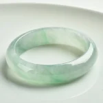 Natural jade bangle bracelet with light green translucent finish, displayed on white ceramic plate showing polished smooth jade surface and natural color variations