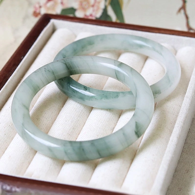Pair of matching natural jade bangles with dark green flower patterns stored in wooden jewelry box with velvet tray, elegant presentation