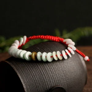 Genuine jade disc bracelet with knotted red rope on textured ceramic container, featuring multi-color jade beads and traditional protective string