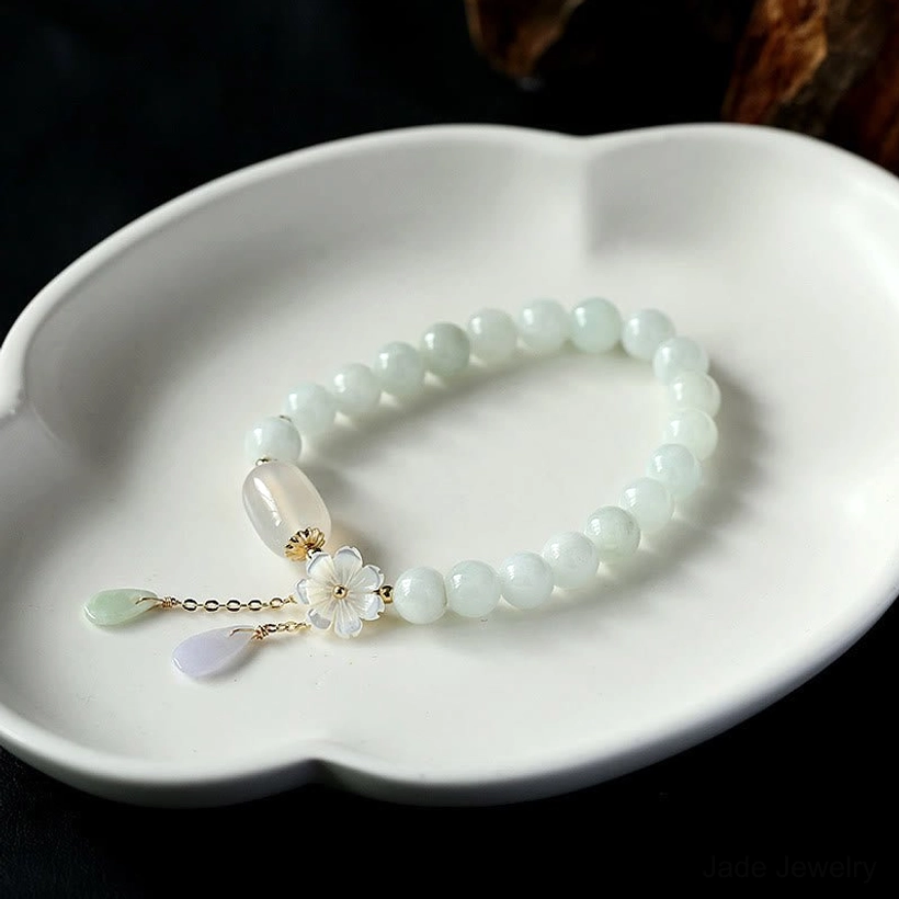 Pale green jade bracelet with polished beads, shell flower charm, and mixed stone pendants on ceramic tray