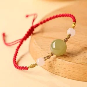 Lucky jade protection bracelet 3/4 angle view showing central green jade bead with red braided cord on wooden coaster