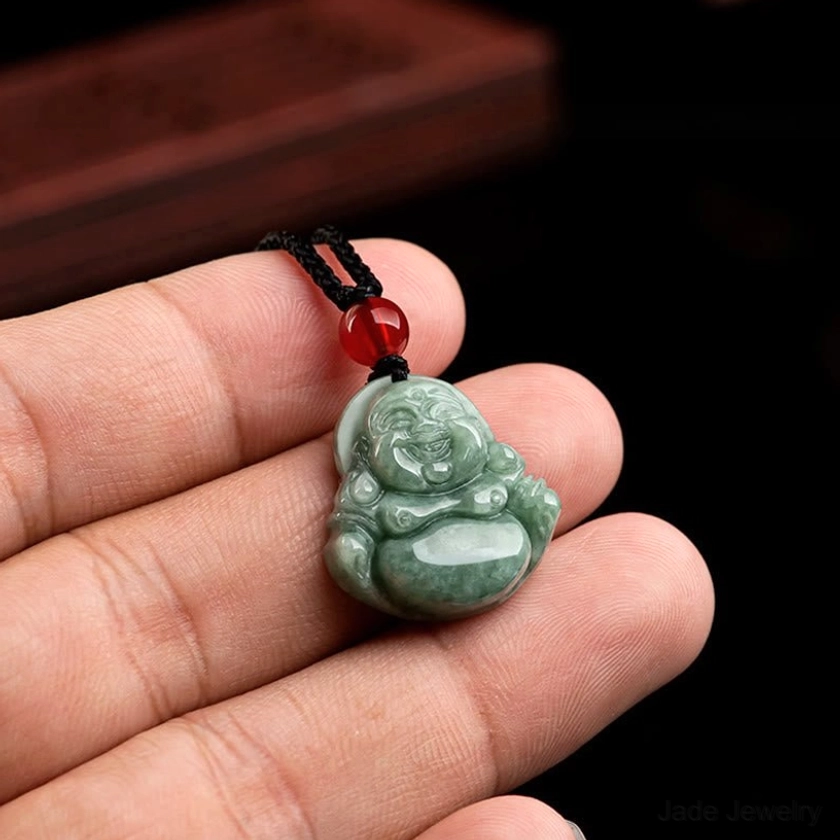 Miniature jade Laughing Buddha pendant held in fingertips showing compact portable size, black braided cord with red bead accent