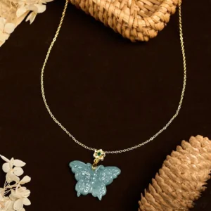 Hand-carved turquoise jade butterfly pendant with delicate gold cable chain against woven rattan decor, overhead angled view