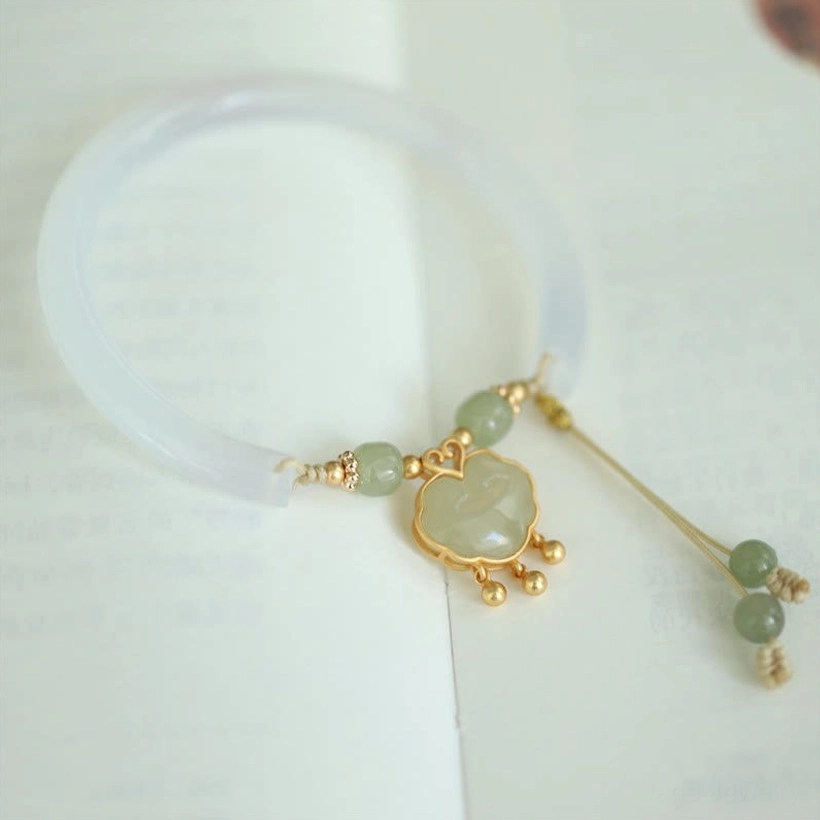 Oblique flat lay of jade ruyi lock bracelet on light paper background, highlighting jade ruyi pendant and tassel accents with gold hardware