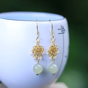 Side view of jade dangle earrings with gold filigree flower charm and pearl accents resting on ceramic cup with green backdrop
