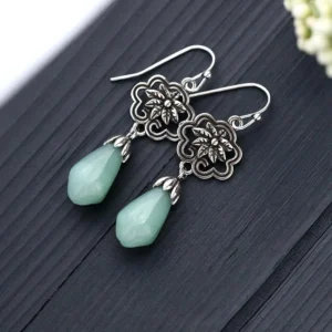 Pair of jade dangle earrings flat lay showing polished pale green jade pendants with silver filigree magnolia flower accents on wood surface