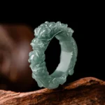 Natural jade dragon ring with intricate hand-carved dragon design, displayed on weathered wood surface showing deep teal jade green color and detailed dragon carving