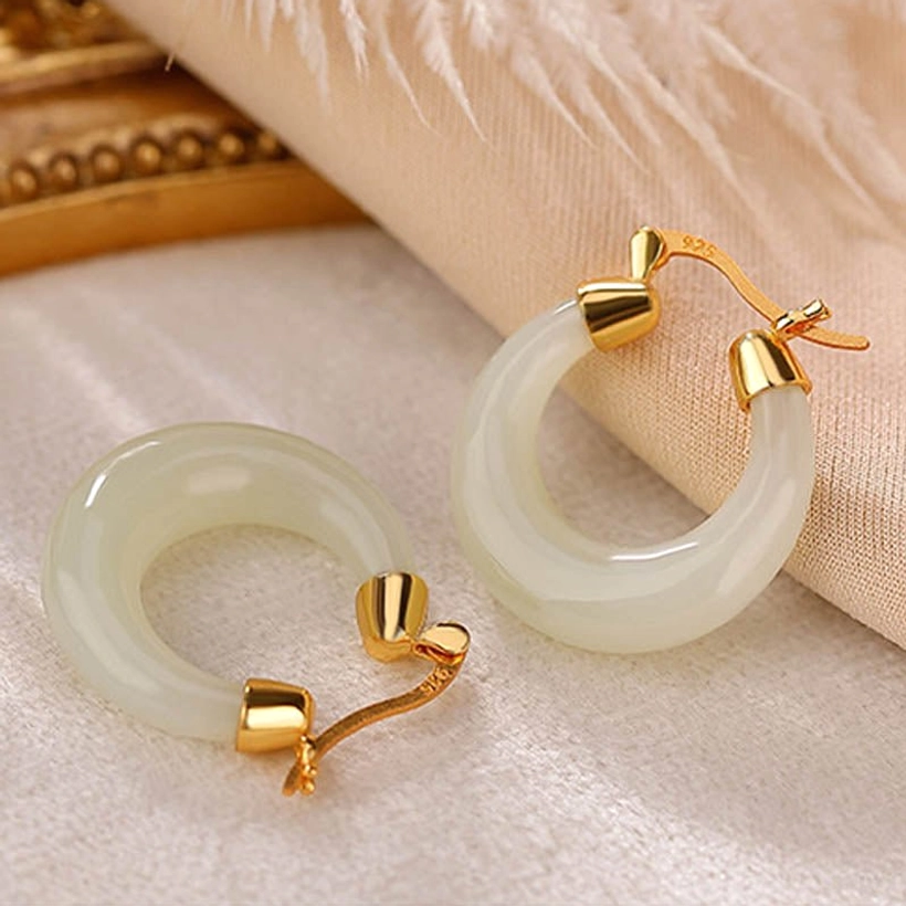 Pair of natural jade drop earrings with one lever-back closure open on satin fabric with golden decor and feather, showing secure fastening mechanism