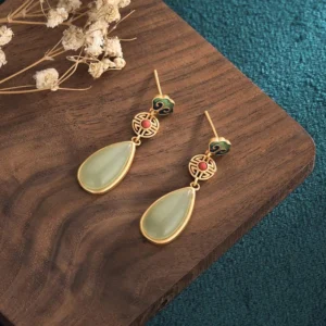 Jade prosperity earrings overhead view on wooden coaster with dried florals, showing hollowed lucky charm design and light green jade drops