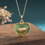 Natural jade elephant pendant necklace with gold-plated copper scalloped frame and polished green jade inlay, displayed on wooden table with dried floral decoration