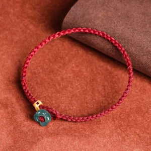Top-down view of jade flower lucky red string bracelet with braided rope, gold metal bead, and dark green jade charm on brown suede backdrop