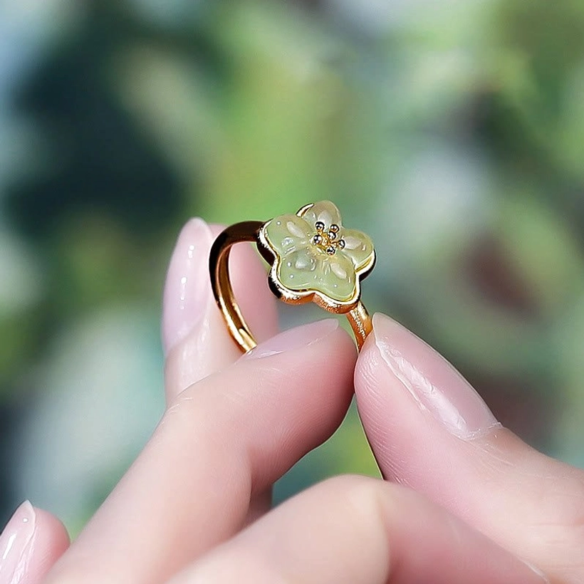 Plum Blossom Jade Ring - Close-up detail of carved flower charm held delicately between fingertips with blurred floral backdrop