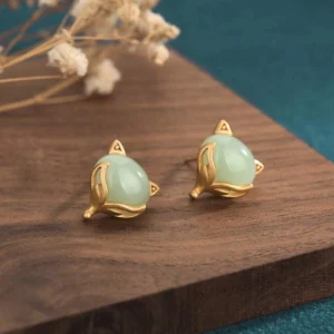 Jade fox stud earrings with light green natural jade stone and gold copper setting, front view flat lay on wooden surface with dried flowers