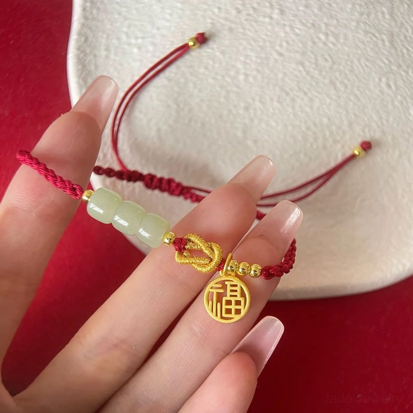 Close-up of handcrafted red rope jade bracelet held in fingers showing light green jade beads and gold decorative knots with Fu charm