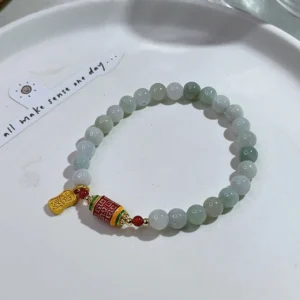 Jade prosperity bracelet flat lay on white plate, showcasing gradient light jade beads, enamel mantra bead, and gold pendant charm for abundance