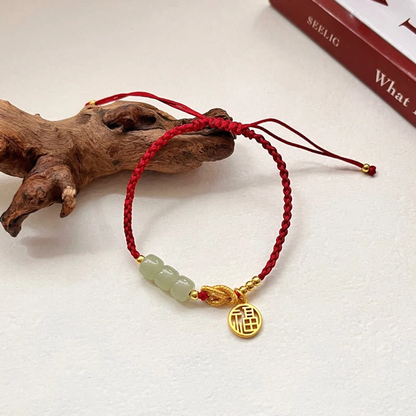 Handcrafted red rope jade Fu character bracelet with gold charm displayed on wooden surface - overhead view of natural jade beads and braided cord