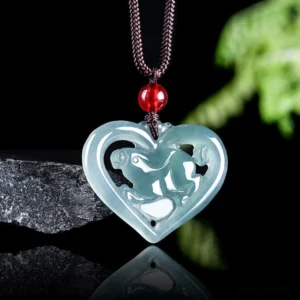 Polished light blue jade heart-shaped rabbit pendant displayed from front overhead view against blurred green foliage backdrop, featuring brown cord with red glass bead
