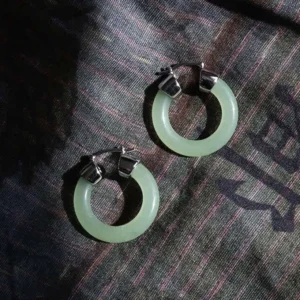 Pale green jade hoop earrings top-down view with silver hardware on dark striped fabric backdrop
