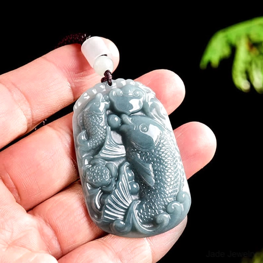 Double koi fish jade pendant held in palm hand-held close-up view, displaying carved koi detail and scale with beaded black cord against nature background