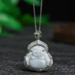 Pale light-green jade Laughing Buddha pendant necklace with silver filigree setting, displayed overhead on dark desktop with green leaf decor