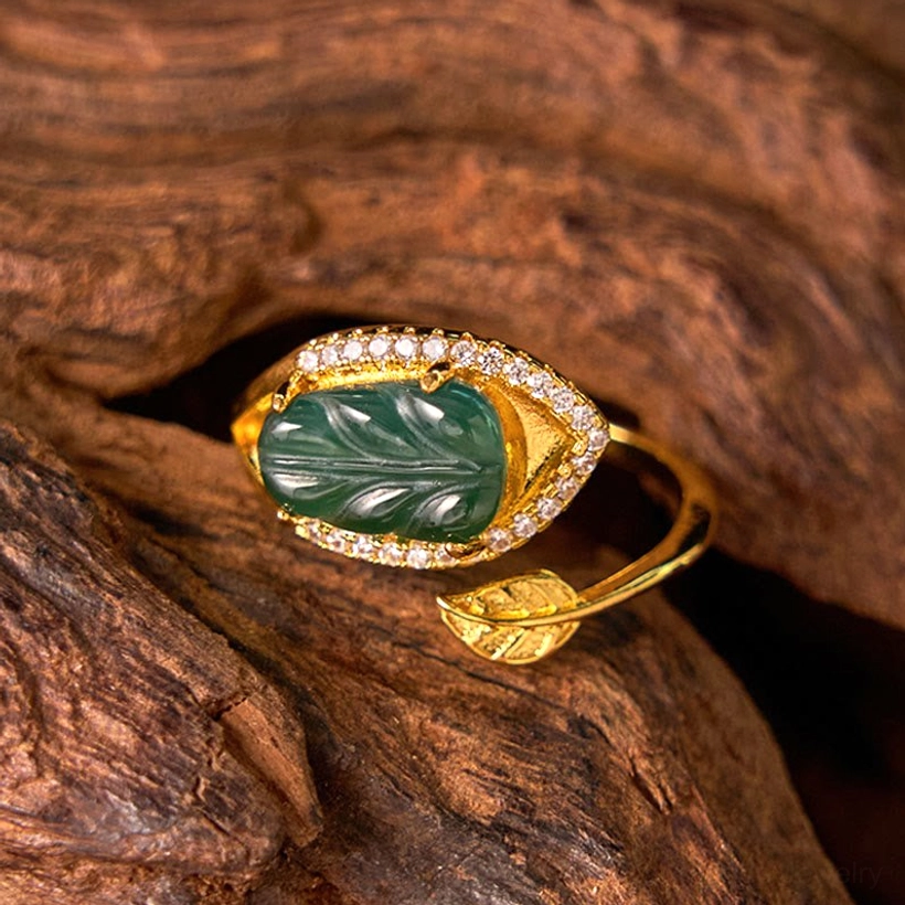 Gold-plated jade leaf adjustable ring - front three-quarter view displaying the decorative leaf ornament on lower band and full crystal halo setting on weathered wood background