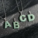 Collection of personalized jade letter pendants A, B, C, D displayed on dark countertop with titanium steel curb chains