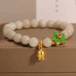 Natural jade lily of the valley luck bracelet three-quarter close-up view with off-white jade beads, green four-leaf flower, and gold lily pendant on brown display