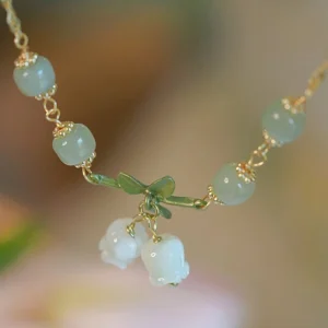 Top-down flat lay close-up of green jade lily of the valley bracelet with cylindrical jade beads, gold metal chain, and enamel lily charms