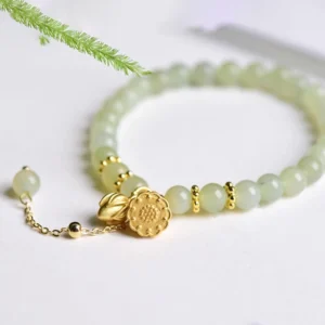 Pale green natural jade beads bracelet with gold lotus charm flat lay on white background with pine branch