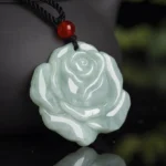 Light green hand-carved jade lotus rose pendant with black braided cord and red agate bead, overhead close-up view on reflective background