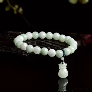 Natural jade money bag charm bracelet with pale green beads displayed on dark glossy surface with reflection, showing side oblique angle view