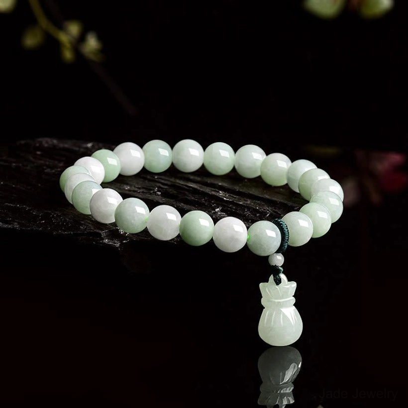 Natural jade money bag charm bracelet with pale green beads displayed on dark glossy surface with reflection, showing side oblique angle view