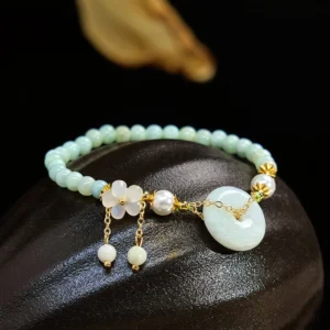 Handcrafted jade peace buckle bracelet with light green natural jade beads, gold hardware, and flower charm displayed on polished black stone