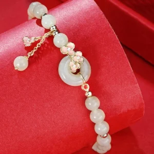 Close-up oblique view of jade peace buckle bracelet with pink enamel flower charms and dangling jade pendant on red fabric background