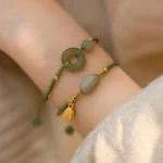 Two green jade peace buckle and lotus braided bracelets worn on forearm with beige knitted sweater, showing adjustable tassel cords