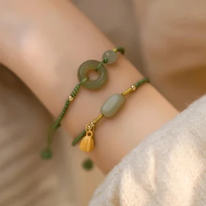 Two green jade peace buckle and lotus braided bracelets worn on forearm with beige knitted sweater, showing adjustable tassel cords