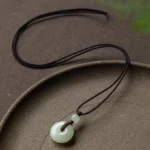 Natural jade peace buckle necklace with dark brown woven cord displayed on ceramic tray showing celadon green jade pendant