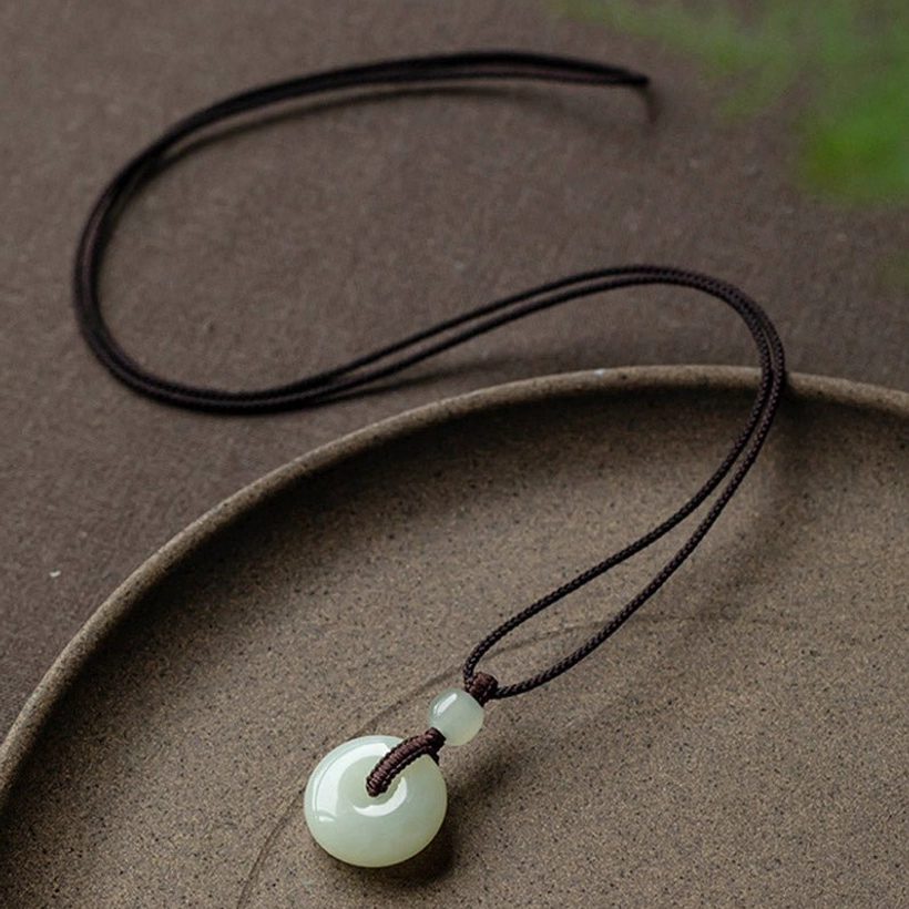Natural jade peace buckle necklace with dark brown woven cord displayed on ceramic tray showing celadon green jade pendant
