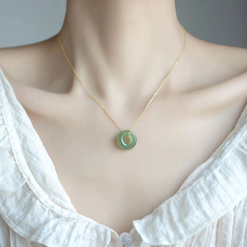 Woman wearing Hetian jade peace buckle necklace at collarbone with delicate gold chain on white linen blouse, styled close-up view