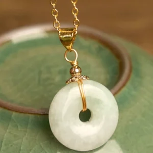 Close-up detail of jade peace buckle pendant showing ornate gold filigree decorative bale with tiny bead accent, smooth polished light green jade ring against green ceramic background