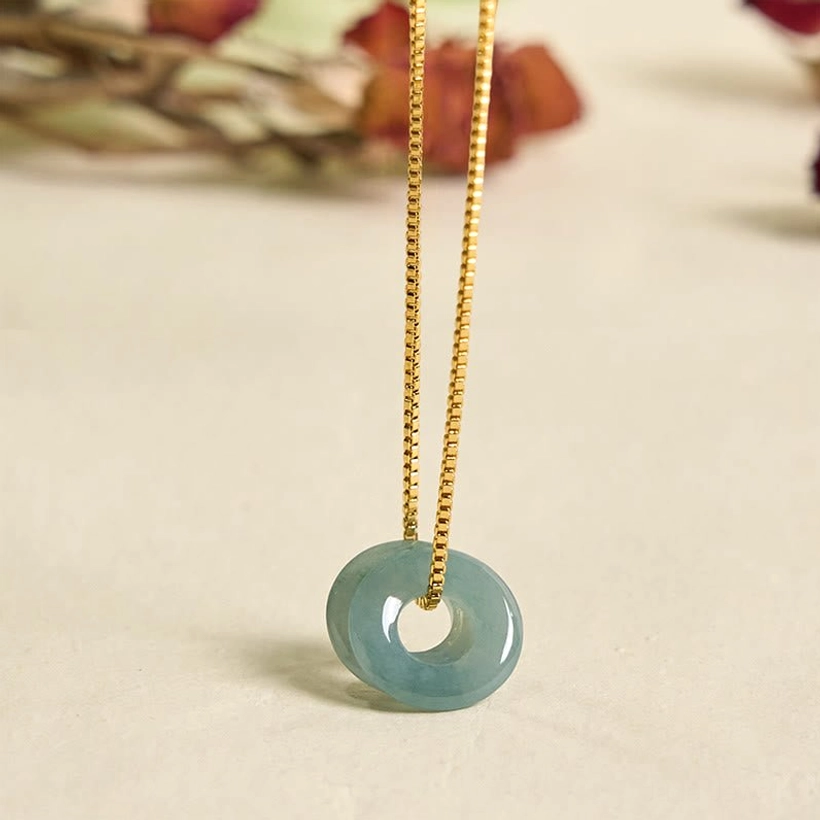 Single jade peace buckle pendant on thin gold chain, vertical close-up on floral wooden table backdrop