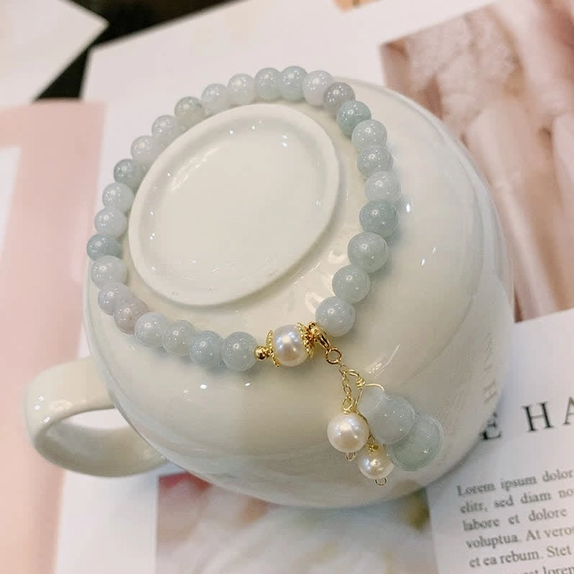Natural jade pearl gourd bracelet flat lay on white ceramic mug showing round pale green jade beads, gold hardware, and pearl gourd charm