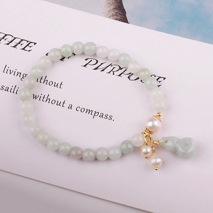 Overhead flat lay of natural jade pearl gourd luck bracelet on book page showing smooth jade beads, gold fittings, and pearl gourd pendant